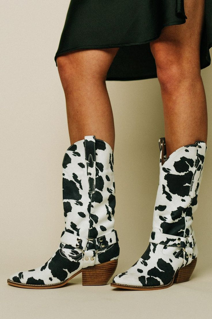 Women's Western cow print boots with a stylish belt accent and pointed toe, perfect for a cowgirl look.